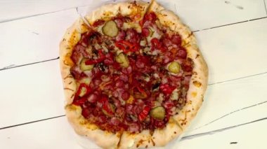 Etli sosisli, biberli, salatalıklı, peynirli dilimlenmiş pizza beyaz ahşap masada döner. Lezzetli ev yapımı sağlıksız fast food. Üst Manzara. Kapat..