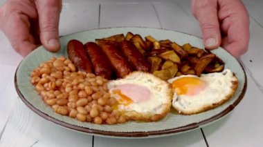 İnsan elleri İngiliz kahvaltısını kızarmış yumurta, sosis, fasulye ve patatesle birlikte ahşap masaya koyar. Ağır çekim. Kapat..