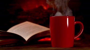 Steaming cup of hot beverage coffee or tea on an wooden board by burning fireplace and open book. Steam rises from a cup. Copy space. Close up.