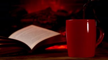 Hot beverage coffee or tea poured into red cup on an wooden board by burning fireplace, and open book.