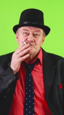 Eski moda, takım elbiseli, sigara içen, şapkasıyla selam veren olgun bir adam. Yaşlı erkek selam vermek için şapkasını kaldırır, sigarasını tüttürür ve dumanı dışarı üfler. Krom anahtar, yeşil ekran. Dikey.