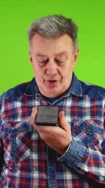 Akıllı telefon tutan ve hoparlörden sesli mesaj kaydeden yaşlı bir adam. Sosyal medya iletişimi, ses komutları, mesajlaşma için akıllı teknolojiler kullanmak. Yeşil ekran, krom anahtar. Dikey.