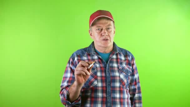 赤い野球の老人はタバコを吸い カメラを見ている タバコタバコを吸う中毒者が目の前で煙を吹き飛ばしている グリーン スクリーン クロマキー — ストック動画