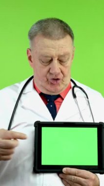 Beyaz önlüklü kıdemli doktor konuşuyor ve yeşil ekranlı tableti işaret ediyor. Uzman sağlık çalışanları danışmanlık yapar ya da tıbbi sunum yapar. Yeşil ekran, krom anahtar. Dikey.