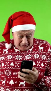 Noel Baba şapkalı ve Xmas kazaklı son sınıf öğrencisi akıllı telefondan dolar kazanıyor. Olgun bir insan para kazanmaktan memnun, başarısından memnun. Yeşil ekran, krom anahtar. Dikey.