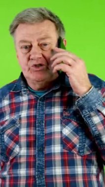 Günlük kıyafetlerle yürüyen ve akıllı telefonuyla konuşan kıdemli bir adam. Modern insanların hayatlarıyla iletişim kurma teknolojisi. Yeşil ekran. Krom anahtar. Dikey.