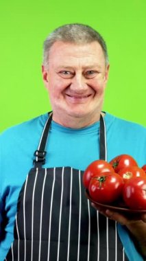 Önlüklü yaşlı aşçı domatesleri işaret ederek sağlıklı yaşam tarzını onaylıyor. Olgun domatesleri işaret eden, vejetaryenlik ya da vejetaryenlik reklamları yapan aşçı. Yeşil ekran, krom anahtar.