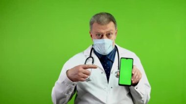 Tıbbi maskeli doktor telefonu yeşil ekranlı tutuyor ve işaret ediyor. Olgun sıhhiyeci, akıllı telefonunda tıbbi haberler ya da reklamlar gösteriyor. Yeşil ekran. Krom anahtar.
