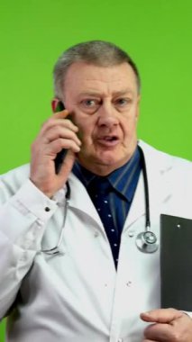 Beyaz önlüklü kıdemli doktor elinde dosyayla yürüyor ve cep telefonuyla konuşuyor. Olgun bir doktor cep telefonuyla yürür ve iletişim kurar ya da danışmanlık yapar. Yeşil ekran. Krom anahtar. Dikey.