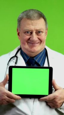 Gülümseyen kıdemli doktor tıbbi haberler veya reklamlar için dijital tablet tutuyor. Olgun sıhhiyeci sosyal medyada yeni tıbbi teknolojilerin veya ilaçların sunumunu yapar. Yeşil ekran. Krom anahtar. Dikey.