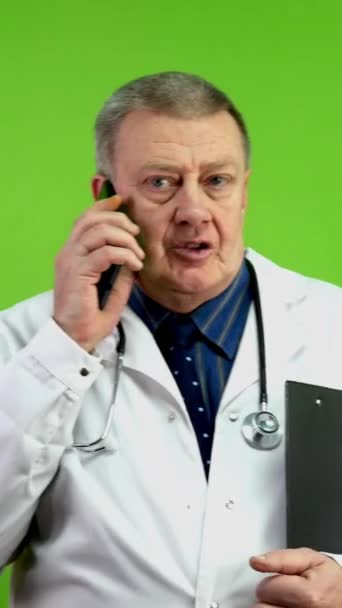 ホワイトコートのシニアドクターは 手のフォルダで歩き 携帯電話で話しています 成熟した医師は歩き回り 携帯電話で通信したり 相談を行います グリーン スクリーン クロマキー バーティカル — ストック動画