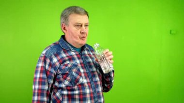 Yaşlı adam susamış ve şeffaf plastik şişeden su içiyor. Normal giyinmiş olgun bir insan susuzluğunu giderir, yorgun ve sıcaktır. Yeşil ekran, krom anahtar.