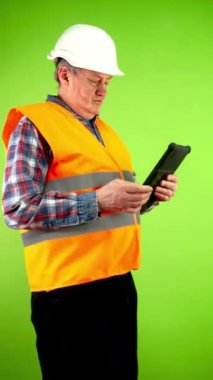 Kıdemli inşaatçı tablet ekranda dokunuyor, tıklıyor, tıklıyor, tıkırdıyor, kayıyor, kaydırıyor. İnşaatta dijital cihaz kullanan olgun bir müteahhit. Yaşayan insanların teknolojisi. Yeşil ekran. Krom anahtar. Dikey.