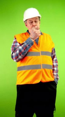 Miğferli kıdemli müteahhit sigara içiyor, rahatlıyor ve duman üflüyor. Olgun işçi zevkle tütün içiyor, bağımlılığı var. Yeşil ekran. Krom anahtar.