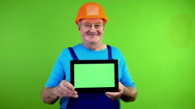 Tulumlu tamirci yatay yeşil ekranlı tablet gösteriyor ve gülümsüyor. Reklam veya pazarlama için elinde dijital tablet bulunan kişi. Yeşil ekran. Krom anahtar.