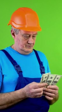 Tulum giymiş, dolar sayıp cebine koyan kıdemli inşaat işçisi. Olgun işçi karı hesaplar ve onaylar. Yeşil ekran. Krom anahtar. Dikey.