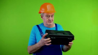 Tulum giyen işçi alet çantasına bakar ve kaliteleriyle övünür. Olgun tamirci alet çantasından memnundur ve baş parmağını kaldırır. Yeşil ekran. Krom anahtar.