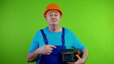 Önlüklü kıdemli gülen işçi aletini gösteriyor ve beni seçiyor, en iyisi benim. Kendisiyle ve işiyle gurur duyan olgun bir tamirci. Tatmin olmuş ve kendinden emin. Yeşil Ekran. Krom anahtar.