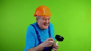 Tulum giymiş kıdemli bir işçi gülüyor ve işaret parmağıyla birini işaret ediyor. Elinde tuvalet pompası olan, dalga geçen, gülen ya da şaka yapan olgun bir tamirci. Yeşil ekran, krom anahtar.