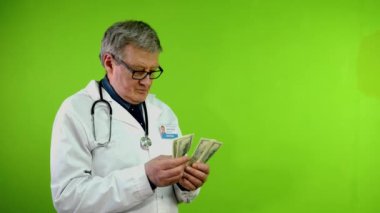 Senior doctor counts money dollars in hands for medical services or bribe received, puts in pocket, gestures shh secret. Green screen, chroma key.