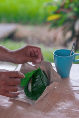 Hand is opening the nasi lemak wrapped in banana leaf with brown paper.