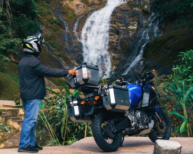 Perak, Malezya - 15 Eylül 2022 Başlık takan bir motorcu motosikletinin yanındaki Lata Kinjang şelalesinde fotoğraf çekiyor..