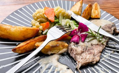 Juicy beef steak with salad and potato wedges along with pepper corn sauce served on black and white stripe on a wooden stone table. Top view