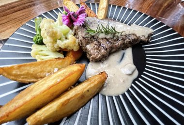 Juicy beef steak with salad and potato wedges along with pepper corn sauce served on black and white stripe on a wooden stone table. Top view