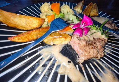 Juicy beef steak with salad and potato wedges along with pepper corn sauce served on black and white stripe on a wooden stone table. Top view
