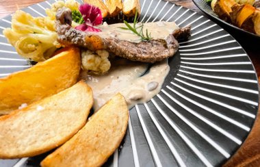 Juicy beef steak with salad and potato wedges along with pepper corn sauce served on black and white stripe on a wooden stone table. Top view