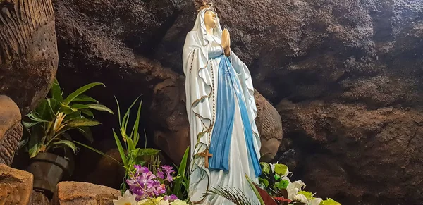 stock image Statue of Holy Virgin Mary in Roman Catholic Church, in the cave of virgin mary, in a rock cave chapel Catholic Church with tropical flowers around in the asian