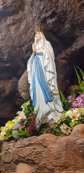 stock image Statue of Holy Virgin Mary in Roman Catholic Church, in the cave of virgin mary, in a rock cave chapel Catholic Church with tropical flowers around in the asian