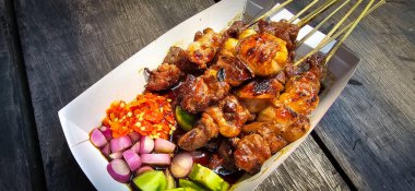Izgara satay, barbekü sosu ve soğan, acı biber ve salatalık turşusu ile servis edilir. Ahşap masanın üstünde seçici odak fotoğrafı.