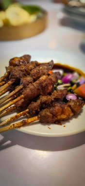 Kuzu eti, ızgara şiş. Soya soslu, soğanlı, domatesli, salatalıklı ve kırmızı biberli, beyaz tabakta servis edilmiş.