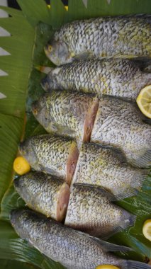 Tam porsiyon Guorami balığı süpermarkette pişirmek ya da satmak için hazırlanan bir çeşit tilapia balığı muz yapraklarıyla güzelce düzenlenir ve limonlarla süslenir, bu kurulum deniz ürünlerini, tropikal mutfağı ve taze yemek malzemelerini ön plana çıkarır..