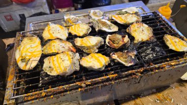 Sokak satıcısı, Tayland pazarlarının hareketli atmosferini gözler önüne sererek, yetenekli ve leziz ızgara istiridye hazırlamak için meşale alesi kullanıyor. Yemek tarifi kitabı için iyi.