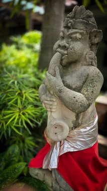 Sakin bir tropikal bahçede bulunan bahçe cücesi geleneksel taş heykeli, çevrenin doğal güzelliğiyle uyumlu canlı yeşillikler ve sanatsal dekorlarla çevrili. Arkaplan için iyi