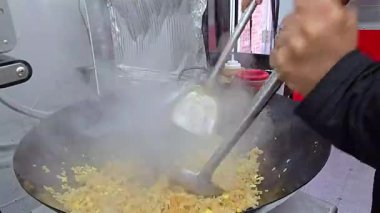 Şef büyük bir tavada pilavı aktif olarak karıştırıyor, profesyonel yemek teknikleri gösteriyor ve mutfak ortamında dumanı üstünde hazırlık yapıyor. Otantik bir aşçılık becerisi ve verimliliği örneği.