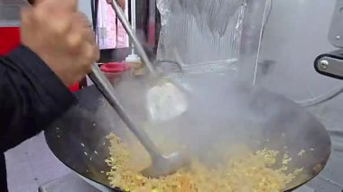 Şef büyük bir tavada pilavı aktif olarak karıştırıyor, profesyonel yemek teknikleri gösteriyor ve mutfak ortamında dumanı üstünde hazırlık yapıyor. Otantik bir aşçılık becerisi ve verimliliği örneği.