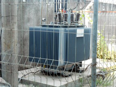 Yüksek voltajlı Transformatör, endüstriyel altyapı ve güvenlik ölçüsünü vurgulayarak endüstriyel bir ortamda güvenli bir çitle korunan yüksek voltajlı elektrik trafosunu tasvir eden bir görüntü.