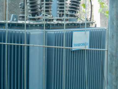 Yüksek voltajlı Transformatör, endüstriyel altyapı ve güvenlik ölçüsünü vurgulayarak endüstriyel bir ortamda güvenli bir çitle korunan yüksek voltajlı elektrik trafosunu tasvir eden bir görüntü.