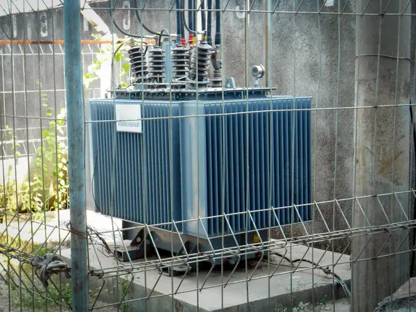 Yüksek voltajlı Transformatör, endüstriyel altyapı ve güvenlik ölçüsünü vurgulayarak endüstriyel bir ortamda güvenli bir çitle korunan yüksek voltajlı elektrik trafosunu tasvir eden bir görüntü.