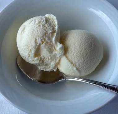 Zarif bir gümüş kaşık eşliğinde yumuşak kumaş yüzeyinde, beyaz bir kasede iki top kremalı vanilyalı dondurma, yumuşak bir kumaş yüzeyinde temiz beyaz bir seramik kasede servis edilir.