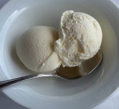 Zarif bir gümüş kaşık eşliğinde yumuşak kumaş yüzeyinde, beyaz bir kasede iki top kremalı vanilyalı dondurma, yumuşak bir kumaş yüzeyinde temiz beyaz bir seramik kasede servis edilir.