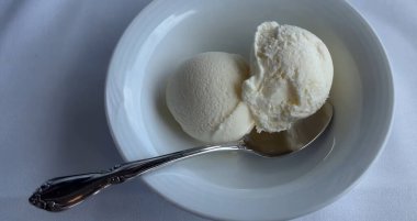 Zarif bir gümüş kaşık eşliğinde yumuşak kumaş yüzeyinde, beyaz bir kasede iki top kremalı vanilyalı dondurma, yumuşak bir kumaş yüzeyinde temiz beyaz bir seramik kasede servis edilir.