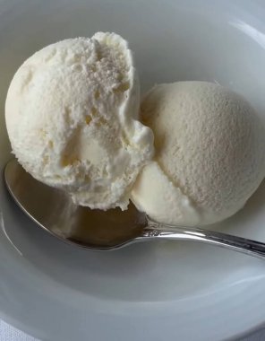 Zarif bir gümüş kaşık eşliğinde yumuşak kumaş yüzeyinde, beyaz bir kasede iki top kremalı vanilyalı dondurma, yumuşak bir kumaş yüzeyinde temiz beyaz bir seramik kasede servis edilir.