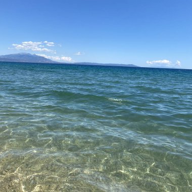 Deniz dalgaları, uzakta bir dağ ve mavi gökyüzünde beyaz bulutlar..