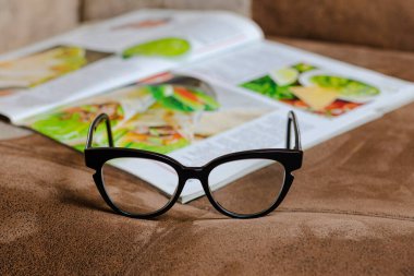 Kanepede siyah çerçeveli kadın gözlükleri, arka planda yemek tarifleri olan bir dergi.