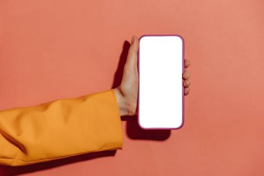 Sarı ceketli bir kadın elinde pembe arka planda beyaz ekran modeli olan bir cep telefonu tutuyor.