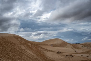 Bulutlu bir gökyüzü olan çöl manzarası. Gökyüzü çoğunlukla bulutlu ve mavi lekeler var. Arka planda kum tepeleri var ve birkaç kişi uzaktan görülebiliyor.